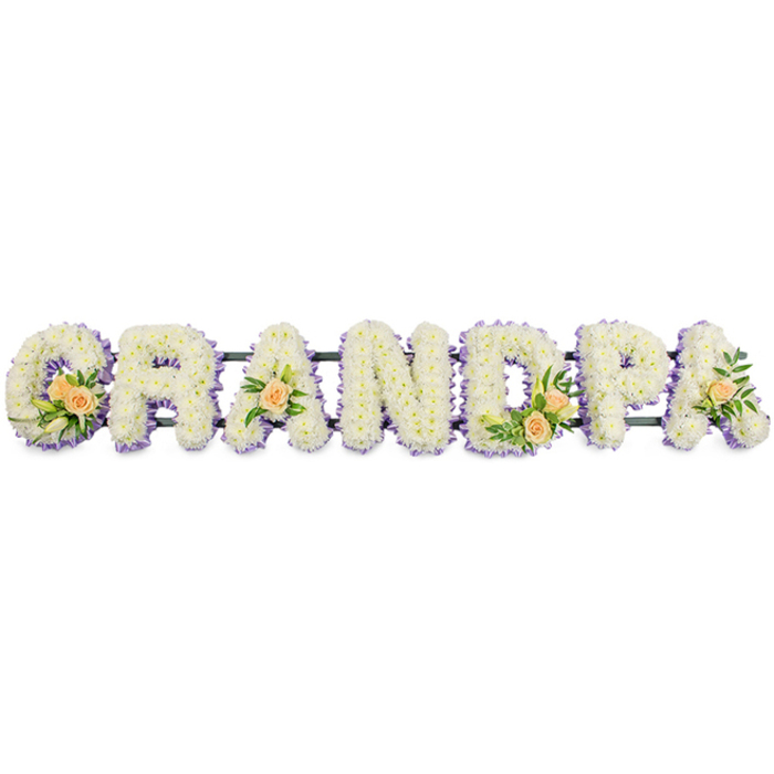 In this carefully composed funeral flower display, the word GRANDPA is spelled out in large, freestanding letters arranged in a straight horizontal line, as if ready to be placed beside a coffin or at the front of a chapel. Each letter is formed on a discreet dark frame and completely encrusted with closely set white chrysanthemums or similar small-headed flowers, so that no gaps in the foliage are visible. The blossoms overlap to create a thick, plush texture, catching the light and giving the word a soft, comforting appearance against the stark white backdrop. Delicately tracing the outer edges of every letter is a border of pale lavender or soft purple blooms, fine enough not to dominate yet strong enough to define each shape-the inner curl of the G, the diagonal legs of the R, and the familiar open triangles of the A's. This subtle contrast of white and lilac suggests calmness, spiritual grace, and a respectful gentleness. To introduce warmth and personal character, several letters are accented with clusters of peach roses and greenery. The G holds the largest grouping, with three or four roses in varying stages of opening, enhanced by glossy green leaves and a few slender white lily buds that extend gracefully above the surface. The first A, the D, and the final A each support a smaller, matching cluster, giving a sense of rhythm along the tribute. This bespoke design could easily be imagined resting in a quiet corner of an Epping churchyard or arriving from a trusted local florist serving homes around Theydon Bois and Loughton, a heartfelt expression of love and remembrance for a treasured grandfather.