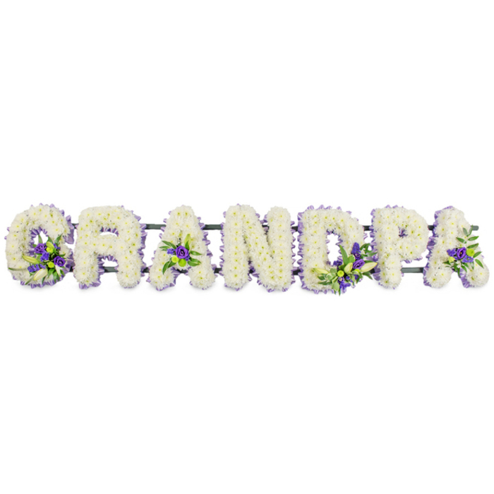 A long, horizontal floral display spells out the word GRANDPA in large block capitals, evoking the presence and steadiness of a beloved grandfather remembered in Epping. Each individual letter is formed on a hidden frame and then lavishly covered with small, tight-headed white flowers-likely chrysanthemums or carnations-so closely set that they create a continuous cushion of soft texture. The white petals appear clean and pristine, the overall effect calm and composed, like a blanket of fresh snow spread evenly over each letter. Within the otherwise white surface, the florist has woven in restrained accents of rich purple and soft lavender blooms, partnered with glossy green foliage. These accents are placed purposefully: a spray curves from the lower edge of the G, another nestles inside the triangular heart of the first A, more flowers glow within the D and the P, and a final touch adorns the last A, bringing the design into gentle visual balance. Around the edges of each letter, a narrow lilac ribbon subtly frames the shape, giving a defined outline without overpowering the simplicity of the white flowers. A dark green bar runs quietly underneath, connecting the letters so the tribute can be displayed as a single piece at a funeral, crematorium, or graveside service near Epping Forest or one of the local churches. The overall mood is tender and composed, offering a personal and respectful way for family members to say goodbye, to stand together and look upon the tribute while recalling stories, familiar sayings, and everyday moments shared with their grandfather.