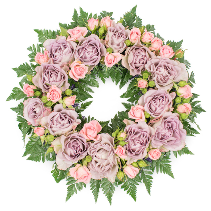 Captured from above against a clean white background, this floral wreath appears as a perfect, unbroken circle of sympathy flowers, its structure both simple and richly detailed. The outer edge is defined by a cushion of dark, glossy green fern fronds, their serrated, feather-like leaves radiating outward to frame the design with a natural, woodland texture. Nestled within this ring of foliage sit numerous large roses in a harmonious blend of soft lavender, muted mauve, and grey-purple tones, each bloom fully open, their many layers of petals curling gently at the tips like fine fabric. Interwoven among these statement roses are delicate clusters of blush pink spray roses and tiny rosebuds, some just beginning to break from their green sepals. These smaller blooms introduce a lightness and tender contrast, softening the more sophisticated tones of the larger flowers and adding rhythm around the wreath. Tiny hints of darker greenery and discreet filler buds are placed between the petals, enriching the depth and creating subtle shadows that make the arrangement feel three-dimensional even on a flat plane. The overall composition is symmetrical and carefully balanced, suggesting the work of an experienced florist used to creating funeral wreaths for services across Epping, from quiet residential streets to gatherings near Epping Forest. The colour palette and form evoke a mood of quiet reverence, ideal for laying at a graveside or memorial, expressing love, gratitude, and respectful farewell without overwhelming the senses. In the stillness of the white background, the wreath seems to hold a moment of calm, offering gentle comfort through its fresh scent and tranquil beauty.