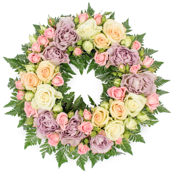 Shown against a bright, uncluttered white background is a perfectly round funeral wreath, abundant with roses and greenery that together create a gentle yet dignified tribute. The wreath's ring is completely filled, forming a rich band of blossoms that flows seamlessly from one bloom to the next, a style often used for memorials in Epping's local churches and crematorium services. Large, open roses in creamy white, soft ivory, and pale peach dominate the design, their petals arranged in concentric layers that curl slightly at the edges, giving a sense of softness you can almost feel. Nestled between these are striking dusty lavender and muted mauve roses, adding depth and a hint of old-world romance, as if the arrangement had been inspired by vintage gardens along the lanes leading towards Epping Forest. Tiny clusters of blush pink spray roses and pale green buds break up the larger forms, introducing finer details and a delicate, almost lace-like texture. The entire circle rests on a foundation of dark green fern fronds, their arching, feathered leaves framing each flower and drawing a crisp outline against the white background. Subtle lighting reveals gentle highlights on the petals and deeper shadows in the recesses of each bloom, creating a sense of volume and quiet stillness. Crafted with care by local florists serving Epping families, this wreath is visually peaceful yet emotionally expressive, ideal for honouring a loved one's life and offering comfort at a time of farewell. It embodies remembrance, continuity, and the enduring bond between the departed and those who remain.
