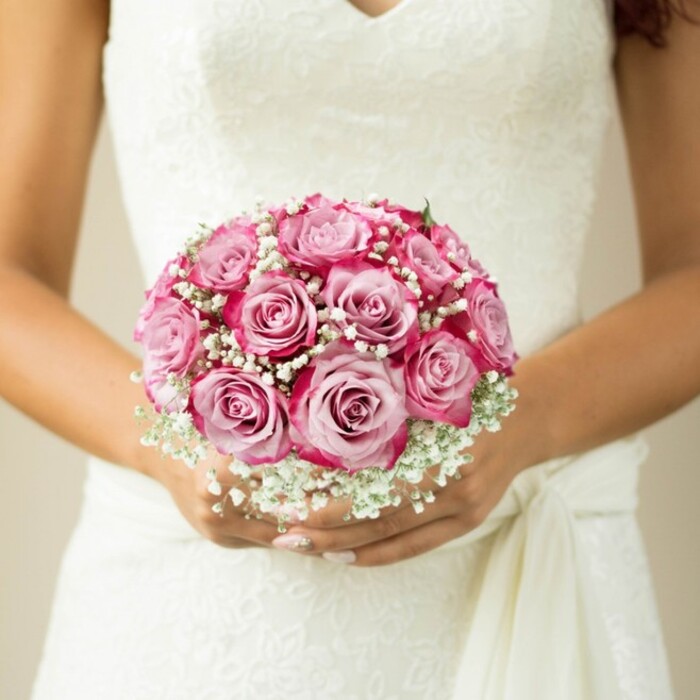 Captured just steps from the bustle of Epping's historic town centre, this close-up scene is one of quintessential bridal romance. The image draws the eye to a flawlessly round bouquet held gracefully against the textured, lace bodice of a classic white wedding dress, open in an elegant V-neck that hints at both modernity and heritage. At the bouquet's heart, a symphony of roses reveals subtle artistry: each petal unfurls from a pale, blushing centre into vibrant magenta or fuchsia outer rims, their colour echoing the wildflowers that bloom along Stonards Hill in late spring. Baby's breath, or gypsophila, froths generously between the roses, creating a diaphanous, cloud-like effect that softens the arrangement's outlines and enhances the bouquet's depth. Barely a wisp of green peeks through, emphasizing the clean, refined shape local brides favour for forest-side ceremonies. The bride's understated, pale-pink manicured nails and the subtle shimmer of her ring finger peek through below the florals, echoing the gentle glamour found in Epping's charming venues. A softly tied sash drapes across the right side, lending a further tactile dimension. The blurred, neutral background ensures that every detail-from petal to lace-is front and centre, making this a bouquet as meaningful and beautiful as a sunrise walk through Epping Forest.