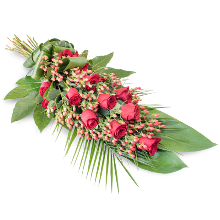 A dignified, elongated floral spray designed for funerals and memorial services is shown resting on a spotless white surface, the neutral backdrop emphasizing the richness of its colours and the precision of its craftsmanship. Along the full length of the tribute, deep red roses are carefully placed, some still in bud, others beginning to unfold into velvety spirals, and a few fully open with layered petals that suggest both strength and tenderness. Interwoven among these roses are rounded clusters of pinkish-red Hypericum berries, their polished surfaces catching the light and offering a softer, slightly coral accent that enhances the palette without overpowering the classic red tones. The flowers are cradled in a structured base of dark green foliage; broad, glossy leaves-similar to Ruscus or Salal-overlap one another to form a dense, protective bed supporting the blooms. From the edges of this base, long, airy palm fronds and fine greenery extend outward, particularly towards the lower right, creating a sweeping, fluid outline that gives the arrangement a sense of movement, as if it were gently cascading. Subtle, slender stems add vertical interest and texture, revealing the detailed layering that a local Epping florist would bring to a bespoke farewell tribute. At the upper left, the stems are neatly aligned and bound with a wide wrap of folded green leaf, providing a smooth, tailored handle and a refined finishing touch. The overall impression is of a carefully considered, heartfelt sympathy arrangement, perfect for honouring a loved one at services held in Epping churches, crematoria, or at quieter family gatherings along the leafy lanes and countryside beyond the town centre.