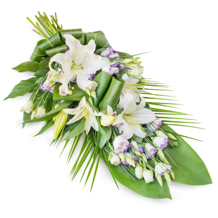 An elegant white and green funeral arrangement, styled as an elongated casket spray or sheaf, is shown diagonally against a bright white backdrop, emphasizing its sculpted outline and layers of texture. At the centre of the tribute are multiple white lilies, likely oriental, their long petals arching outward in star-shaped blooms. Some flowers are fully open, revealing creamy throats and soft yellow stamens dusted with pollen, while others remain in partially open or closed bud form, slender and poised, symbolizing hope, remembrance, and the continuation of family stories. Around them, clusters of white lisianthus (Eustoma) bring a softer, more intricate detail; their gently ruffled petals are edged with touches of lavender or muted purple, adding a whisper of colour that never disrupts the calm, monochrome mood. Many lisianthus buds remain tightly curled, like tiny scrolls, giving the eye subtle points of interest along the length of the spray. The composition widens near the top left where the stems are gathered, then narrows to a refined point toward the bottom right, the overall shape reminiscent of a falling leaf or tender teardrop. Supporting this floral heart is a rich bed of greenery. Broad, dark green leaves anchor the design, some left flat to create a strong backdrop, others expertly folded and rolled to introduce sculptural accents that suggest deliberate, caring craftsmanship. Long, slender palm-style fronds emerge from the right-hand side, sweeping faintly outward and downward to extend the line of the spray, contributing to its sense of gentle movement. The finished piece conveys quiet dignity and peace, ideal for funeral services, memorial gatherings, or farewell ceremonies in Epping's churches and chapels, offering a graceful floral expression of sympathy and respect.