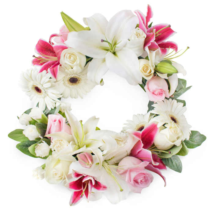 A lush circular funeral wreath sits against a plain white background, its full ring of flowers forming a tranquil, contemplative tribute that would look at home in any Epping memorial setting. The arrangement is built around large white lilies, spaced evenly around the circle so their broad, satin-like petals and prominent golden stamens become the main focal points. Between them are bold accents of deep pink and fuchsia lilies, whose star-shaped blooms and darker, speckled throats lend the wreath a sophisticated richness and gentle contrast. White gerbera daisies are dotted throughout, their crisp petals and contrasting dark centres adding both structure and a soft, hopeful brightness. Clusters of blush-pink roses appear in various stages of opening, from tight, pointed buds to partially unfurled blooms, symbolising tenderness and the unfolding of cherished memories. Creamy white roses, more fully opened with layered, spiralling petals, contribute to the wreath's depth and classical elegance. Smaller white blossoms-likely miniature carnations or spray roses-fill in between the larger flowers, thickening the circle and creating a plush, almost cushion-like texture. Underneath, and occasionally peeking through, is an abundance of fresh green foliage in varied shapes and tones, providing a natural, verdant backdrop that frames the colours beautifully. The wreath's open centre emphasises the circular symbolism of eternity and continuity, making it a fitting floral tribute for funerals, celebrations of life, or remembrance services held in and around Epping, from quiet churchyards to gatherings near the edges of Epping Forest.