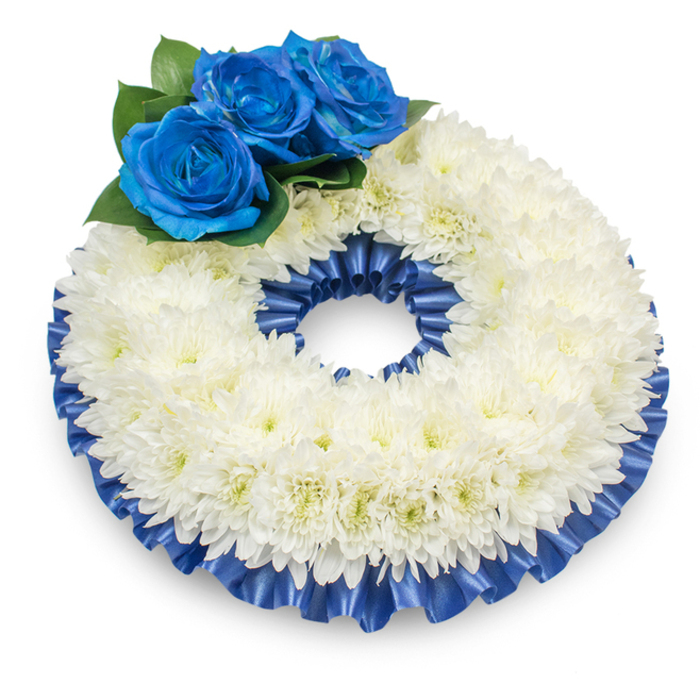 Viewed from a modestly raised angle against a spotless white background, this circular memorial wreath reads as a composed, elegant statement of condolence. The principal mass is formed from densely packed white chrysanthemums, their soft, feathery petals creating a slightly domed, cushion-like ring whose texture invites a moment of quiet attention; faint creamy centres show through in the light, lending a subtle warmth. At the upper left, three intense electric-blue roses form a distinct focal cluster-their richly pigmented, velvety petals unfurl in compact spirals and are cushioned by dark green foliage that accentuates the depth of colour. A pleated satin ribbon in matching deep cobalt traces both the inner and outer edges of the chrysanthemum ring, its glossy folds catching highlights and offering a refined frame that defines the wreath's silhouette. The arrangement rests in a soft pool of shadow on the white plane, giving the composition depth and a gentle sense of gravity appropriate to a memorial context. As an experienced local florist, I picture this piece placed near a chapel doorway or on a viewing table for services around the Market Square or along Station Road in Epping-an arrangement that balances tradition and personal sentiment. The blue and white palette conveys both dignity and a quiet comfort; it reads as a calm, noble tribute intended to honour memory, express condolences, and provide a reassuring visual anchor for those assembled to remember a loved one.