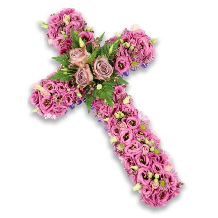 This image shows a beautifully detailed floral cross tribute, set crisply against a bright white background that emphasizes every petal and curve of the arrangement. The cross itself is three-dimensional and generously proportioned, its entire surface carpeted with fresh blossoms in a harmonious blend of soft pink, mauve, lilac, and delicate purple. At the intersection of the horizontal and vertical arms, three larger roses in a muted dusty rose to light mauve tone form a clear focal point, their fully opened heads revealing whorls of petals that suggest tenderness and enduring affection. These central roses are embraced by sprays of rich green foliage, including fine fern fronds and compact leaves, the greenery providing a gentle frame and anchoring the pastel shades with natural depth. Stretching along the arms and down the length of the cross, ranks of lisianthus blossoms are massed together, their trumpet-shaped, slightly ruffled petals overlapping to create a soft yet structured texture. Some lisianthus blooms display darker, more pronounced centers, adding visual contrast amidst the lighter tones. Threaded throughout, clusters of minuscule pink and purple filler flowers, pale green buds, and discrete white blossoms offer delicate points of interest, inviting a closer look. Just at the borders of the design, especially along the sides of the arms, fine lines of darker purple ribbon or a fabric base can be glimpsed beneath the flowers, giving the arrangement a polished, expertly finished feel. The cross conveys a sense of peace, reverence, and love, the sort of thoughtful funeral tribute families in Epping might arrange for a service at a local church or crematorium, where this handcrafted piece can quietly express sympathy and honor a cherished life.