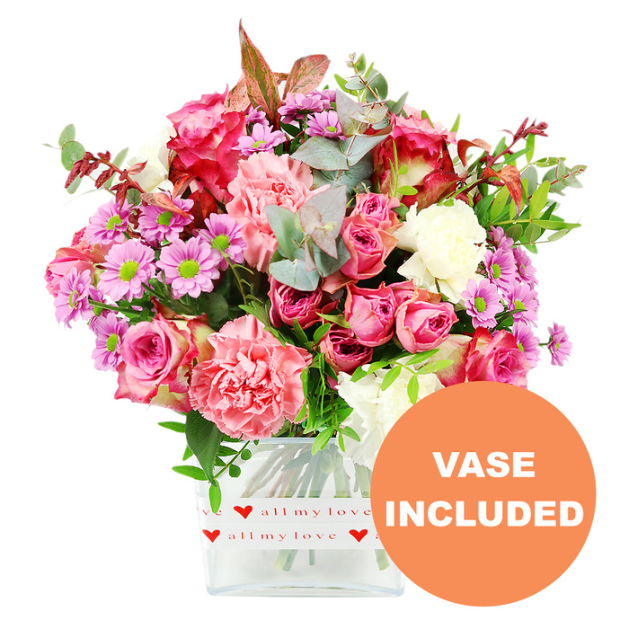 Against a clean, uncluttered white background, a richly textured bouquet in a clear rectangular glass vase appears as though it has just been placed on a sunlit table in an Epping home. The arrangement is dominated by roses and carnations in an array of pinks, from deep fuchsia to delicate pastel blush, interwoven so that each shade gently plays off the next. The roses, some fully open and others still unfurling, show layered, velvety petals that curl softly outwards, while the carnations bring clusters of frilly, ruffled petals in coral and peach-pink tones that create volume and softness. Scattered among these larger blooms are small daisy-like chrysanthemums in lavender and purple with bright green centres, their cheerful faces lifting the whole palette. Creamy white blooms are carefully tucked between the colours, giving balance and creating the sense of a well-composed piece designed by an experienced local florist. Surrounding the flowers is a generous collar of foliage: rounded eucalyptus leaves in soft silvery green, darker glossy greenery, and the occasional reddish-brown leaf that hints at late-summer or early-autumn warmth, like the changing shades you might notice on a stroll near Epping Forest. The stems are neatly aligned inside the transparent vase, which has softly rounded corners. Around its lower portion, a white decorative band printed with repeating red hearts and the phrase all my love in flowing script clearly sets a romantic, affectionate tone. In the lower right corner of the photograph, an orange circular overlay carries the words VASE INCLUDED in bold white capitals, signalling that this is a complete, ready-to-display floral gift. The overall impression is of a carefully crafted bouquet, ideal for anniversary celebrations, birthdays, or thoughtful same-day delivery to addresses across Epping and the surrounding villages.