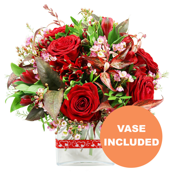 Set against a spotless white background, this opulent red bouquet forms a perfect, rounded dome of blooms, as though a piece of midsummer warmth has been gathered and placed in a vase. Dominating the composition, several large red roses stand proudly, their petals dense and velvety, some still tightly coiled while others unfurl into full, layered rosettes that suggest romance and devotion. Woven around them are clusters of deep red flowers akin to alstroemeria and carnations, each stem offering multiple smaller blossoms that add a sense of fullness and rhythm, like a chorus supporting a soloist. Throughout the bouquet, airy sprigs of pale pink and white waxflower are carefully threaded, their tiny blossoms and bead-like buds creating a fine, lacy texture that softens the intensity of the reds. The flowers are embraced by a rich frame of greenery: broad, glossy leaves, lighter green shoots, and striking foliage in burgundy and reddish-brown tones, some with delicate veins that catch the light and lend a contemporary, almost artistic flair. This lush arrangement is anchored in a sleek, clear rectangular glass vase with gently rounded edges, allowing a glimpse of the stems and water within and giving the piece a polished, modern character that would sit beautifully in an Epping living room or a cosy café near the High Street. Around the vase, a red ribbon patterned with white heart outlines is tied neatly, instantly evoking themes of love, anniversaries, and Valentine's surprises. In the bottom right corner of the image, a bold orange circular overlay with clean white uppercase lettering reads VASE INCLUDED, making it visually clear that this romantic, ready-to-display arrangement is delivered as shown, complete and effortless for the recipient.