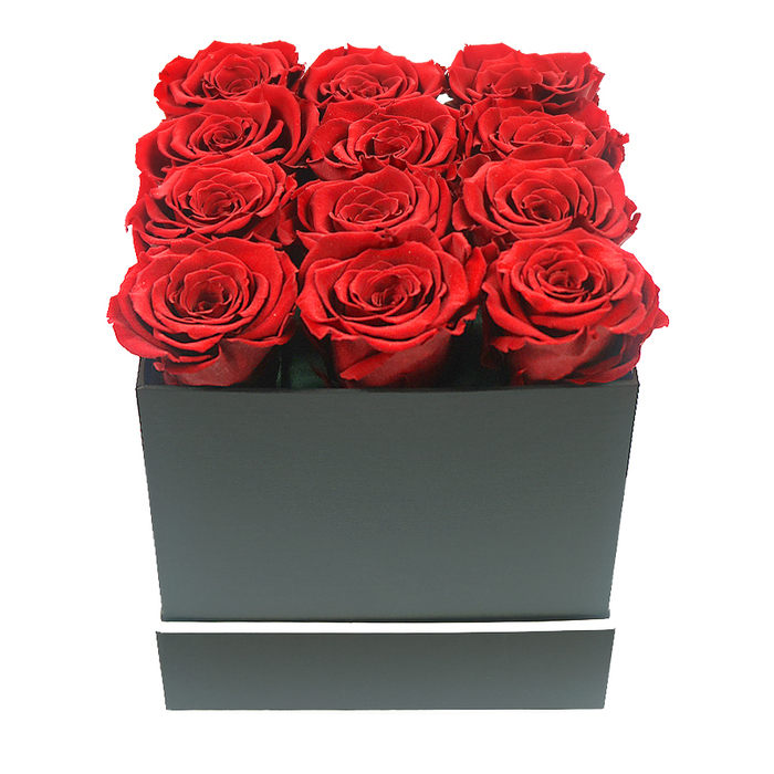 The photograph shows a meticulously crafted rose arrangement that feels both dramatic and serene, the kind of piece often requested for significant occasions across Epping. Dominating the frame is a perfectly square, matte black box, its sides sharp and unembellished except for a slim white stripe that runs horizontally around the lower section like a tailored trim on a designer coat. Nestled at the top, twelve luxurious red roses are arranged in a neat 3-by-4 pattern, filling the entire opening so completely that no interior of the box is visible. Each rose is fully open and carefully shaped, with densely layered petals that appear soft and velvety, their colour a deep, consistent crimson with gentle tonal shifts where the light brushes the petal edges. The flower heads project slightly above the rim of the box, creating a tactile, cushion-like surface of blooms that you can easily imagine brushing with your fingertips. The scene is lit with soft, diffused lighting that avoids harsh glare, instead caressing the curves of each petal and tracing delicate shadows into the heart of the roses and along the box's corners. The backdrop is an unblemished white, free of props or clutter, so the arrangement seems to float in space, its rich reds and strong blacks standing out with quiet intensity. The overall impression is of carefully curated luxury and enduring affection, ideal for a special Valentine's gesture in Ivy Chimneys, a proposal celebration in Theydon Mount, or a sophisticated centrepiece for a dinner table near Stonards Hill. Designed in the elegant spirit that local clients associate with Flowers Epping, this preserved rose box suggests love that remains vibrant long after the day has passed.