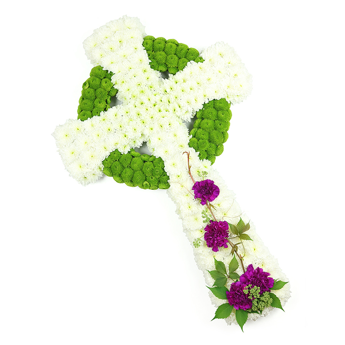 This photograph shows a diagonally positioned Celtic cross funeral arrangement, stretching from the top left corner of the frame towards the bottom right, set against a bright white studio backdrop that makes every colour and texture stand out. The structure of the cross is densely packed with pristine white chrysanthemum blooms, each small flower head nestled closely against the next to create a soft, cushioned effect across the upright stem and outstretched arms. The chrysanthemums' tiny petals form a consistent, plush texture, evoking serenity and quiet reverence, much like the stillness around St John's Church in Epping early in the morning. Encircling the centre of the cross and tracing the distinctive Celtic halo is a ring of lime-green button chrysanthemums. These rounded, tightly formed blooms draw a clear circular shape where the vertical and horizontal arms meet, while additional bands and pockets of the same bright green flowers highlight the inner knot pattern and corners, giving the design visual depth and a hand-carved, stone-like feel translated into petals. On the lower vertical arm, three carefully composed groups of deep purple carnations introduce a dramatic contrast. The carnations are lush and layered, their velvety petals curling inwards, suggesting tenderness and remembrance. They are interlaced with small sprigs of dark green foliage and slender, twisting stems that trail lightly across the white chrysanthemum base, reminiscent of ivy climbing old gravestones in the quieter parts of Epping Cemetery. The lighting is clean and even, enhancing the freshness of the flowers and the artisan skill involved. This Celtic cross tribute would be a poignant choice for a funeral or memorial service in Epping, reflecting faith, heritage, and the enduring bond with a loved one who has passed.