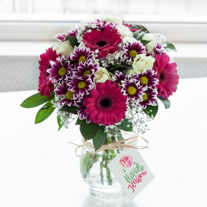 A beautifully crafted mixed flower arrangement is captured in soft natural light, standing in a squat, clear glass vase with subtle vertical texture, set against a bright, gently blurred white backdrop reminiscent of a sunlit window in an Epping townhouse. Dominating the bouquet are several generous gerbera daisies in rich fuchsia and magenta shades, their petals arranged in neat concentric layers around dark central disks that feel almost velvet-black, giving the arrangement a bold, contemporary heart. Clustered around them are multi-petaled chrysanthemums, their daisy-like blooms displaying elegant purple tips that gradually fade into white, each flower finishing in a bright yellow centre that echoes the warmth of afternoon sun over Epping High Street. Interlaced between these statement flowers are creamy white spray roses; some are fully open, revealing swirling soft petals, while others are still tight buds, suggesting new beginnings and lending a gentle, romantic touch. Airy sprays of baby's breath thread through the bouquet, their tiny white blossoms creating a cloud-like effect and softening the strong pink and purple tones. Deep green foliage fans outward and upward from the base, giving structure and a sense of freshness, as if the stems were gathered only moments ago. The clear vase reveals neatly trimmed stems submerged in clean water, and at its neck a length of rustic twine is tied in a casual bow, supporting a small, white rectangular tag decorated with a simple pink rose motif and flowing script. The design feels like a joyful, thoughtful gift-a bouquet you might have delivered by Flowers Epping to a family in Theydon Bois, to celebrate a birthday, a promotion, or even just to brighten a quiet corner after a walk along the edge of Epping Forest, bringing colour, texture, and a sense of affectionate celebration into the home.