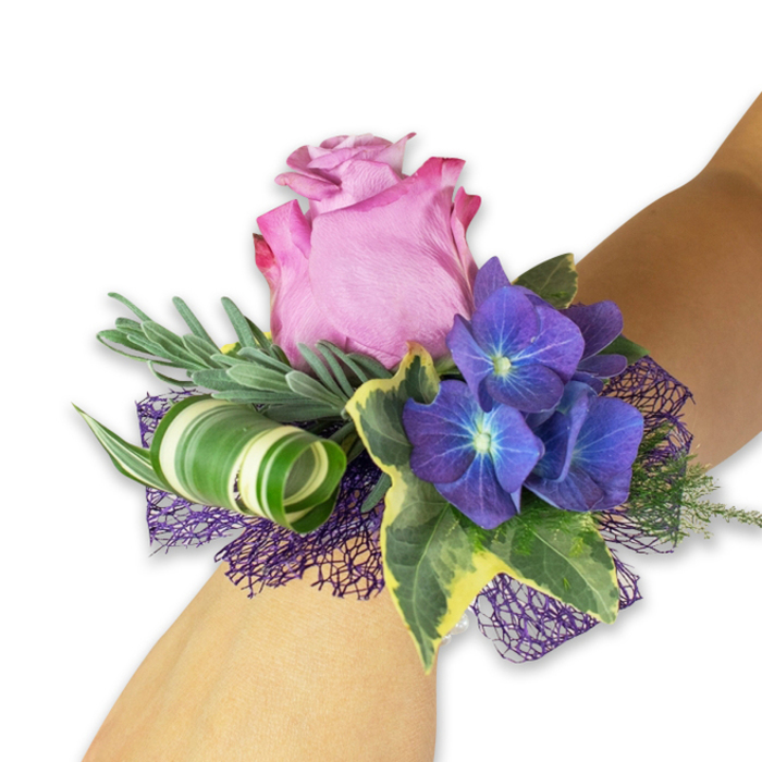 On a fair-skinned wrist, a hand-crafted corsage combines classic romance with contemporary detail, photographed against a crisp white backdrop that makes each colour sing. The focal bloom is a large pink rose, partially open so its concentric petals reveal a subtle ombré-from a richer fuchsia at the outer ruffle to a pale blush toward the centre-inviting touch and releasing a gentle, floral scent. Counterbalancing the rose are three prominent clusters of deep indigo hydrangea; their compact florets carry tiny blue-tinted centres that glint like small stars within the purple mass, providing texture and depth. Variegated ivy leaves arch gracefully, their dark green cores rimmed in creamy yellow, contributing an elegant, traditional frame to the composition. Behind the flowers, slender, silvery-green stems-suggestive of eucalyptus or lavender-give a soft, airy backdrop and a whisper of herbal fragrance. Adding a modern flourish is a broad green leaf striped in white, tightly curled into a sculptural spiral that punctuates the arrangement with a bold linear accent. Below, an open-weave dark purple mesh nestles the blooms, offering a lacy shadow that amplifies the hydrangea's tones and adds lightness to the silhouette. The overall effect reads as sophisticated and wearable: a corsage that would complement bridesmaids walking through the village towards the church or a graduate stepping out from a celebration on Epping Green. It's a fresh, balanced design-textural, tactile, and crafted to feel both personal and memorable for special local occasions.