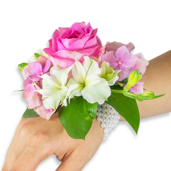This elegant wrist corsage is photographed on a left hand against a clean white background, each element rendered with careful horticultural detail. Dominating the arrangement is a full, hot fuchsia-pink rose whose richly layered petals form a velvety, tactile centre. Surrounding the rose, soft pastel-pink hydrangea florets add plush volume and a light, airy texture; their clustered petals create a gentle backdrop that enhances the rose's intensity. Sprays of crisp white alstroemeria (Peruvian lilies) are interspersed throughout, their pointed petals revealing faint green streaks at the throat and offering bright highlights that energise the palette. To the right, a compact group of pale green alstroemeria buds and a few barely opened pink buds impart a sense of growth and future bloom.
A foundation of broad, glossy emerald leaves frames the flowers and introduces a cooling contrast to the warm pinks. The arrangement is secured with a sophisticated multi-strand bracelet made of small pearlescent white beads, which wraps around the wrist and traces a delicate net across the back of the hand, the pearls reflecting soft studio lighting. Visible skin is a light-to-medium tone, giving scale and intimacy to the image. Designed for formal moments-prom corsages, bridal bouquets' accompanying accessories, or graduation ceremonies-this piece demonstrates refined craftsmanship, balanced proportion, and a whispered scent of rose and fresh greenery. Imagine it worn for a spring wedding near St. John's Road or a reception after a stroll through Epping Forest-subtle, polished, and lovingly assembled for special occasions.