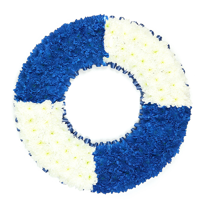 A nautical-themed funeral wreath, meticulously crafted in the unmistakable shape of a lifebuoy, is set against an immaculate white background, allowing its deep blue and white colour story to command full attention. The ring is tightly packed with countless small flowers, most likely fine chrysanthemum heads, creating a smooth yet subtly rippled surface, like gentle waves when viewed from a distance. The wreath is cleanly divided into four equal quarters, alternating in colour: rich royal blue blooms fill the top left and bottom right sections, while pristine white flowers occupy the top right and bottom left. This disciplined pattern gives the tribute a calm, rhythmic balance, reminiscent of traditional maritime safety rings seen at harbours along the coast, even though it is displayed here as if freshly delivered to a funeral home near Epping High Street. Each bloom is placed with care so that the petals overlap, forming a continuous carpet of colour that curves gracefully around the central void. The inner edge of the circle is delicately lined with a slim band of dark blue ribbon, pleated into small, regular folds that add a refined, textile contrast to the softness of the petals. The ribbon also subtly darkens the inner circumference, reinforcing the circular outline and making the central opening stand out in quiet relief. Soft, even lighting ensures that every texture-from the velvety depth of the blue flowers to the cool clarity of the white-remains visible without harsh glare. This lifebuoy wreath feels like a heartfelt expression of safe passage and enduring care, a fitting memorial for someone whose life was intertwined with the sea, with rescue services, or simply with offering steadfast support to family and friends across the Epping community.