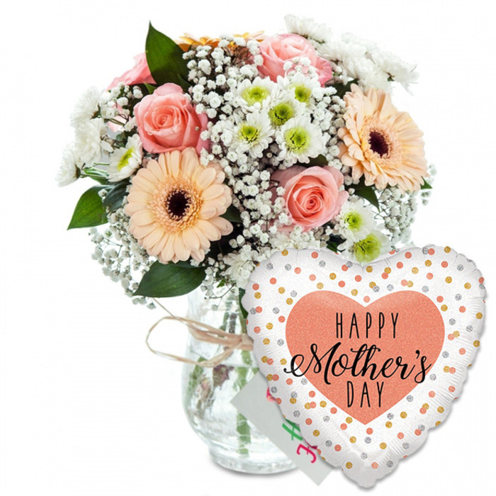A lovingly prepared Mother's Day gift set is displayed against a bright white background, the simplicity of the setting drawing full attention to the colours and emotions carried in the flowers and balloon. On the left, a lush, rounded bouquet sits securely in a clear glass jar-style vase, its transparency revealing a tidy cluster of stems and a hint of water glimmering inside. A length of natural twine is tied around the neck in an easy bow, adding a homely, handcrafted feel that would be at home on a kitchen table in Epping or near Theydon Bois. The bouquet itself is a harmonious blend of soft spring tones: several peach and pale pink roses open fully, their layered petals curling gently outward like satin. Nearby, two prominent peach-toned gerbera daisies with deep brown centres stand out cheerfully, their broad petals forming perfect circles that bring a playful energy. Interspersed between these main blooms are clouds of delicate white baby's breath, lending a light, misty texture, and small daisy-like white chrysanthemums, each with a subtle green eye that introduces a fresh, natural contrast. The flowers are nestled among rich green foliage that frames the composition, giving it depth and the feeling of a freshly picked garden bundle, the sort of arrangement a local florist might prepare for doorstep delivery along Epping High Street or towards Buckhurst Hill. To the lower right, partially overlapping the vase, a heart-shaped foil balloon adds a festive note. Its crisp white surface is scattered with shiny gold and silver polka dots, while a central peach heart holds the greeting HAPPY Mother's DAY in a mix of bold and script black lettering. Just behind the balloon, the corner of a small white greeting card peeks through, subtly reinforcing the theme of gratitude and celebration for mums. The whole ensemble radiates warmth, perfect for marking Mother's Day with a thoughtful gesture anywhere in and around Epping.