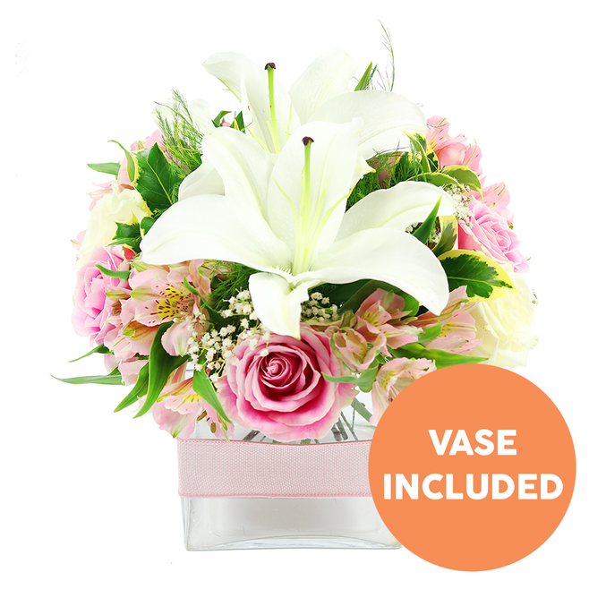 A thoughtfully composed bouquet of white and pink flowers is showcased against a smooth white background, capturing the calm elegance of a fresh flower delivery arriving at a townhouse near Epping Forest or a quiet home off Theydon Road. At its heart, two large white Oriental lilies open generously, each bloom displaying velvety, slightly curved petals that form a star-like silhouette. Deep reddish-brown stamens stand proudly at their centres, adding an earthy contrast to the cool whiteness of the petals. Surrounding these focal blooms, soft pink roses are arranged in a dense cluster, some rose heads still cupped tight, others fully open to reveal intricate spirals of silky petals that suggest romance and warmth. Interspersed throughout are pink and white alstroemeria, their slender petals blushed with pastel tones and peppered with subtle yellow flecks, lending detail and texture. Wisps of white baby's breath (gypsophila) weave through the design like fine mist, lightening the composition and drawing the eye from one bloom to the next. The bouquet rests in a clear, square glass vase, the straight lines of the container giving a modern counterpoint to the soft curves of the flowers. Around the centre of the vase runs a pale pink ribbon, neatly tied and perfectly aligned, adding a gentle, gift-like finish. Beneath the blossoms, a lush bed of greenery-from glossy, rounded leaves to more delicate feathery stems-creates depth and a sense of fullness, suggesting the freshness of a walk through nearby Epping Forest Park. In the lower right corner of the image, a vivid orange circular badge with bold white capital lettering clearly states VASE INCLUDED, indicating that the vase is part of the floral gift. The overall mood is one of quiet sophistication and heartfelt sentiment, suited to birthdays, anniversaries, or simply brightening a loved one's day in Epping.