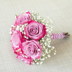 Crafted with artistry in Epping by an experienced local florist, this lush bridal bouquet radiates romance and elegance. The heart of the arrangement is a medley of roses in a striking two-tone palette-mauve and lavender shades glowing within, framed by intense fuchsia and magenta edges. Each rose's petals gently curl outward, creating a rich tapestry of colour and form. Sprigs of snow-white baby's breath weave between the larger roses, lending a light texture and softening touch reminiscent of early morning mists along Epping Forest. Carefully bound by a deep plum satin ribbon, the stems are further enhanced with a spiral of iridescent pearls-each bead reflecting subtle glimmers in the natural light, suggesting careful attention to bridal tradition and detail. The bouquet's base reveals neatly cut, natural green stems for a refreshing, organic finish. Placed atop a light beige, linen-textured surface-perhaps evoking the welcoming interiors of Epping's country homes-the arrangement exudes warmth and approachability, inviting the observer to imagine the fragrance and tactile beauty of freshly gathered blooms. Ideal for weddings in local venues or as a luxurious centrepiece for a milestone celebration, this bouquet from Flowers Epping embodies the grace, care, and vibrant spirit cherished throughout the local community.