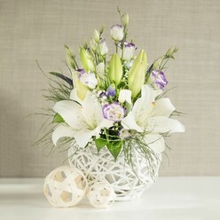 A refined floral centerpiece is captured in gentle, even light, the kind you might find streaming through a dining room window in Epping on a clear day. Center stage is a round white vase with an intricate lattice design, its open-weave texture recalling handwoven rattan or wicker, light and breathable yet visually striking. Rising from this sculptural base is an arrangement of pristine white lilies, their star-shaped blooms unfolding fully, each petal smooth and luminous, with stamens delicately dusted in warm pollen. Around them, elongated green buds promise further blossoms, adding a sense of movement and unfolding time, ideal for a wedding breakfast table or evening reception. Interwoven with the lilies are lisianthus flowers in shades of white and cream, many brushed at the edges with soft lavender-purple, like watercolour strokes. Some are in full bloom, their layered petals creating gentle spirals, while others remain in slender bud, lending a sense of quiet anticipation. Fine, feathery greenery arcs through the design, supported by broader, glossy leaves, creating depth and a lush, garden-inspired silhouette that would feel at home near Epping Forest or along quiet residential streets. Tiny white filler flowers, likely baby's breath, and small touches of deeper violet blooms peek through, adding lightness and delicate contrast. On the smooth white tabletop rest two decorative spheres in pale wicker tones, echoing the vase's pattern and completing the harmonious look. The composition exudes calm elegance, perfectly suited to wedding tables in Epping venues, anniversary dinners, or intimate gatherings where understated beauty and soft, fragrant florals set the mood.