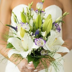 Captured on the eve of a joyous wedding in Epping, this bridal bouquet tells a delicate love story in flowers. Creamy-white Oriental lilies, their fragrance suggesting both celebration and serenity, spread wide under soft, natural light, each petal highlighted by gentle freckles and golden stamens that glimmer like early morning sunbeams over Copped Hall. Accompanying the lilies are frilled white lisianthus blooms edged in whimsical violet, their texture and subtle colour transitioning beautifully between tradition and contemporary taste-a nod to the evolving romance of couples across Epping Lane. Between these larger blossoms, deep purple Veronica stems create visual rhythm and movement, dancing with tufts of baby's breath that evoke the delicate clouds over Epping's Market Square. Fern-like greenery and bold lily leaves frame the bouquet, their fresh dewiness reminiscent of early walks in Epping Forest. Gently held by hands adorned with a soft blush manicure, the bouquet's stems are neatly wrapped, creating a tactile link between the floral artistry and the bride's anticipation. Crafted with a master's touch, this arrangement radiates fresh beginnings and gentle nostalgia, and would feel right at home among the church aisles or grand venues sprinkled across Essex. Every detail, from the colour harmony to the careful layering of textures, reflects a bouquet designed not just to accompany a dress, but to illuminate a day filled with joy, love, and the warm spirit unique to Epping's wedding celebrations.
