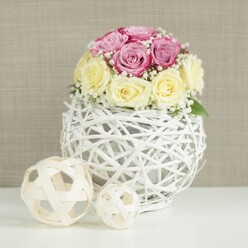 A romantic wedding-style centerpiece is pictured in soft, natural light, showcasing a compact dome of roses that feels both luxurious and understated, much like a carefully curated Epping townhouse interior. About a dozen roses sit closely grouped, their petals overlapping to create a plush, rounded silhouette. The colour palette is soothing and refined: half the blossoms glow in a delicate blush-pink with a subtle lavender undertone, while the rest are a mellow cream-yellow, the kind of gentle shade that evokes candlelight in an evening reception at a venue near Epping Forest. Nestled between these roses are fine wisps of white baby's breath, their tiny blooms catching the light and adding a lace-like texture that softens the overall design. The arrangement rises from a striking white spherical vase, formed from thin, painted branches or wicker woven into an open lattice, like a decorative globe. This airy, handcrafted vessel sits on a pristine white surface, its shadows barely visible, letting the intricate weave speak for itself. To the left of the vase, two decorative wicker or rattan balls in natural wood tones echo the open-weave pattern and lend a relaxed, organic note that would suit a rustic-chic celebration in or around Epping. Behind everything stretches a softly textured beige wall, perhaps linen or a subtle wallpaper, which keeps attention firmly on the flowers. The lighting is calm and even, with no harsh contrasts, making the pastel petals and snowy-white wicker appear especially soft. The entire composition feels like a refined table arrangement ready for a wedding breakfast, an anniversary lunch, or a special delivery from a local florist to quietly transform an everyday dining table into something memorable.
