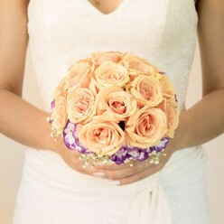 In this intimate, mid-frame photograph, the camera lingers on the torso of a bride standing serenely, her classic ivory gown forming a soft backdrop for a meticulously arranged round bouquet. The fabric of the dress is smooth and gently structured, with delicate lace tracing the bodice and edging a modest V-neckline, suggesting a timeless style so often chosen for weddings in and around Epping's historic churches and countryside venues. Resting in her lightly manicured hands is a sumptuous cluster of roses that forms the rounded heart of the bouquet. Each rose is a study in warm pastel nuance, with petals shifting from pale peach and apricot at the outer edges to slightly deeper, almost coral pink tones at the centre. The flowers are tightly packed to create a compact, domed silhouette, giving a sense of fullness and quiet luxury. Beneath this cushion of roses, a distinctive band of purple ruffled blooms, resembling lisianthus, wraps around the base. Their deep violet centres fade into creamy white at the edges, creating a two-tone effect that frames the roses like a painter's careful border. Interspersed between these blooms are tiny, cloudlike tufts of white baby's breath, lending a light, airy texture and a hint of traditional bridal charm. The bride's thumbs gently meet at the stems, one nail catching the light with a subtle glitter or ombré finish, a small personal flourish amid the classic design. The lighting is soft and even, without harsh shadow, placing full emphasis on the colours and craftsmanship of the bouquet. The arrangement feels perfectly suited to a romantic ceremony near Epping Forest or a reception in a local country house, its peach and purple palette evoking warmth, tenderness, and a promise of lasting memories.