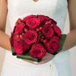Evoking a sense of drama and grace reminiscent of elegant ceremonies held across Epping, this remarkable bridal bouquet is the essence of romance immortalized in scarlet hues. Each rose, from tightly furled buds to luxuriously unfurling petals, is chosen for depth of colour and flawless form, creating a captivating sea of red. Dotted throughout, clusters of gleaming red hypericum berries add both texture and a joyful sparkle, resembling tiny treasures gathered during a stroll through Epping's historic market gardens. The bouquet's base is swathed in wide, glossy green leaves, meticulously folded to frame the blooms and lend structure, reminiscent of the lush foliage seen along Bell Common paths. Cradled with care in the bride's gently clasped hands-manicured in understated elegance-the bouquet sits against a backdrop of delicate lacework and flowing white fabric, details softly illuminated to highlight their artistry. The air is tinged with the imagined scent of fresh roses, grounding the senses in an atmosphere both festive and intimate, reflecting the joy and anticipation present in every Epping wedding. This bouquet, crafted with an eye for classic tradition and contemporary beauty, is not just a visual delight but an enduring symbol-a signature creation often seen at unforgettable wedding receptions throughout the community. Perfect for couples wishing to make a statement of devotion and abundance, it offers a poetic tribute to love's enduring vow.