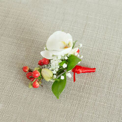 A carefully crafted floral boutonniere lies centered on a lightly woven, beige linen-style fabric, its gentle texture evoking the relaxed, natural charm of homes and wedding barns around Epping and the nearby countryside. The focal point is a flawless white Phalaenopsis orchid, its wide, silky petals opening in a serene, sculptural curve. At the heart of the bloom, a soft wash of creamy yellow and muted orange adds warmth, drawing the eye to the flower's intricate throat. Around this central orchid, minute white blossoms of baby's breath form delicate, cloud-like clusters, giving the piece an airy, almost ethereal veil of texture. Below and to one side, a tight spray of glossy, round red berries introduces a lively burst of colour, reminiscent of winter hedgerows along the lanes leading towards Epping Forest. A few lighter green, bead-like buds nestle among them, suggesting freshness and the promise of new blooms. Smooth, medium-green leaves in varying shapes-some broad and tapered, others slim and elegant-are threaded through the composition, lending depth and a fresh-from-the-garden feel. At the base, the stems are drawn together and wrapped in a vibrant red satin ribbon, tied into a small, refined knot with short tails that fall neatly against the fabric. The lighting is soft and natural, enhancing the contrast between the crisp white petals, the vivid berries, and the calm neutrality of the background. This single, thoughtfully balanced boutonniere feels ideal for a groom or guest at a wedding, prom, or festive celebration in Epping, capturing a sense of quiet luxury and heartfelt attention to detail.
