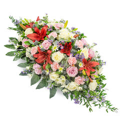 A graceful, double-ended funeral spray is shown against a crisp white background, its colours and textures forming a soothing river of blooms that runs from left to right. The arrangement is generously packed, yet thoughtfully composed, with no harsh edges, only soft, tapering lines created by trailing greenery and carefully angled stems. Dominating the design are deep red stargazer or Oriental lilies, their large, star-shaped petals edged in a richer crimson and dusted with darker speckles, creating a striking focal point. Nestled among them are clusters of cream and soft pink roses, some barely opening, others fully blossomed, their layered petals giving a sense of softness and quiet intimacy, like cherished memories unfolding. Pale pink carnations, with delicately fringed petals, tuck into the gaps, while small white chrysanthemums or daisy-like flowers bring freshness and light into the heart of the spray. Punctuating the warm tones are steel-blue thistles and sprays of lavender-blue filler flowers that dance lightly between the larger heads, adding texture and cool contrast. The entire piece rests on a rich bed of foliage, including broad, deep green leaves and finer, ferny greens that arc gently outward and beyond the flowers, lending movement and a natural, garden-like feel. The mood is one of calm remembrance and gentle farewell, perfectly suited to a service in Epping or a chapel gathering near the forest. As a local florist would explain to a family, this style of tribute combines classic funeral flowers with contemporary touches, creating a comforting expression of sympathy and enduring love.