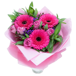 This arrangement reads like a springtime note sent in colour: bold, joyful, and intimately made. Three fuchsia Gerbera daisies dominate the centre with wide, layered petals fanning from their dark, dramatic centres, creating an immediate focal point that feels both playful and confident. Around them, lavender-pink roses sit with petals still tight and velvety, suggesting freshness and a soft romantic restraint. Little clusters of purple statice tuck into the spaces between, their papery texture and cooler tone adding depth and a whisper of wildflower charm. Lush green foliage - broad salal leaves and stems of silvery eucalyptus - builds a generous backdrop, adding weight and a crisp, dewy leaf scent that balances the sweeter florals. The bouquet is carefully wrapped: an inner layer of pale pink matte tissue gives a gentle base, and an outer sheet of sheer, crinkled fuchsia-pink cellophane lends a faint shimmer and modern polish. The rim of a clear glass vase is visible at the bottom, suggesting the arrangement is ready to sit on a kitchen table or greeting corner. Light plays softly across petals so that colours read layered rather than flat, and the overall fragrance is delicate - roses offering a classic perfume while gerberas contribute a fresher, green note. Perfect for celebrating a birthday, saying thank you, or cheering a friend, this bouquet carries the everyday warmth of Epping and nearby lanes such as Bell Common and St. John's Road, and it's suited for local delivery when you need an immediate, sincerely bright gift.