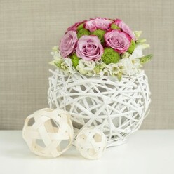 An artfully composed floral centrepiece is captured against a softly blurred beige backdrop, the kind of elegant table arrangement you might see at a wedding reception in one of Epping's country-style venues. At the centre of the image sits a distinctive spherical vase, woven from slender white rattan-like branches into an open, basket-style globe that feels both contemporary and rustic, as if inspired by the natural textures of nearby Epping Forest. Rising from this airy base is a carefully shaped dome of flowers: large, fully open roses in soft pink and muted lavender tones form the main body of the bouquet, each bloom showing layers of velvety petals and subtle hints of deeper pink along the edges. Tucked between the roses are vivid green button chrysanthemums, their compact, rounded heads adding a fresh burst of colour and a pleasing, tactile texture that contrasts with the smooth rose petals. Around the lower edge of the arrangement, small white filler flowers nestle among the greenery, creating a gentle, cloud-like fringe that softens the transition between the florals and the woven vase. On the white tabletop to the left of the vase, two decorative spheres made from pale wicker or wood reinforce the natural, hand-crafted theme; their open, criss-crossed structures echo the vase's design and introduce a relaxed, organic rhythm to the composition. The overall effect is calm and refined, an image that suggests the quiet anticipation of guests arriving, and the care of a local Epping florist arranging each table with precision and a personal touch.