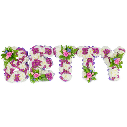 When language feels insufficient, this bespoke floral name piece for BETTY speaks with quiet grace. Each substantial letter is formed on a dense bed of creamy white chrysanthemums whose rounded, snowy blooms feel soft to the touch and provide a serene canvas for the design. Contrasting magenta-purple chrysanthemums trace inner and outer contours, creating a jewel-like definition that reads like a ribbon of colour across the white ground. A thin purple fabric edge around each letter lends a polished silhouette, while clusters of delicate pink roses - some fully open, others in a shy bud stage - are arranged as small posies on the B, at the central bar of the first T and on the Y, their gentle perfume mingling with the green freshness of fern fronds. Interspersed tiny blue flowers, reminiscent of cornflower or delphinium, add cool highlights that catch the eye and lift the composition. The lighting in the image is even and soft, emphasizing the texture of petals and the workmanship of floristry, and the white background keeps attention on the flowers themselves. This personalised tribute is conceived for funerals or memorial gatherings in Epping, whether placed near the altar at a chapel close to Epping station or displayed at a family wake on the High Street; it is a calm, dignified expression of remembrance that comforts as it commemorates. Crafted with attentive technique and a gentle colour harmony, it reads as both a celebration of a life and a tender farewell.