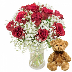 Set against a crisp white background, this image captures a timeless romantic gesture that feels perfectly at home in Epping, whether imagined on a quiet street near the Forest or a cosy house off the High Road. Dominating the left side is a lush arrangement of approximately twelve deep red roses, gathered in a clear glass vase. Each rose is rich and velvety, the petals tightly layered and gently curving outwards, some still in that promising stage before full bloom while others open generously, revealing their full, heart-shaped centres. Soft sprays of white baby's breath weave in and around the roses, like tiny stars scattered across a night sky, creating a delicate contrast that makes the crimson tones appear even deeper and more luxurious. The transparent vase reveals straight green stems and clear water within, offering a glimpse of the florist's careful conditioning and fresh preparation. To the right of the bouquet, a plush light brown teddy bear sits upright, its slightly rounded body and gently drooping paws giving it an inviting, cuddle-ready presence. The fur appears fluffy and touchable, with small black button eyes that lend the bear a kind, almost shy expression. A neat dark brown ribbon is tied in a simple bow at its neck, adding a polished, gift-ready detail. This combination of roses and teddy bear suggests a planned surprise-perhaps delivered by Flowers Epping for a Valentine's breakfast in Coopersale, an anniversary dinner near Epping Station, or a birthday treat that blends luxury flowers with childlike comfort and affection.