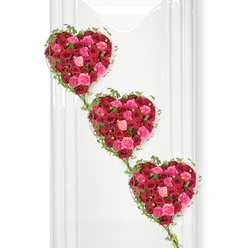 Three opulent hearts made entirely from roses descend gracefully across a pure white backdrop, forming a striking funeral tribute that feels both romantic and reverent, as if designed for a thoughtful farewell at a Epping church or woodland memorial. The hearts are aligned in a soft diagonal, each one slightly offset so they seem to flow downwards like a gentle path of memories. Every heart is densely filled with roses in rich crimson, bright fuchsia, and delicate blush pink, the colours blending into a tapestry of passion and tenderness. The petals overlap in tight, luxurious layers, with some roses fully unfurled and others still cupped, creating a tactile, almost cloud-like texture. Encircling each heart, fine strands of glossy green ivy trace the outline with precision, their small, fresh leaves adding a natural frame that keeps the shapes clear and defined. These ivy tendrils extend from one heart to the next, forming a living link that echoes the continuity of love and shared stories. The bright white wall behind them reflects light onto the flowers, making every shade of red and pink stand out in sharp clarity while subtle shadows lend depth. This floral design feels deeply personal, the kind of bespoke tribute a family in Epping might choose to express love, gratitude, and remembrance. It balances strong emotion with calm beauty, combining classic red roses, softer pink tones, and trailing greenery into a dignified, heartfelt arrangement for a final goodbye.
