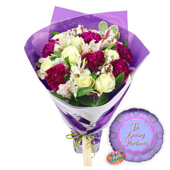 Set against a pure white backdrop, this romantic floral gift set feels like it has just been finished on the workbench of an Epping High Street flower shop, ready to be carried home through the lanes towards Epping Forest. The central focus is a generous hand-tied bouquet, alive with contrast and careful craftsmanship. About a dozen creamy white roses form the calm, elegant core, their tightly coiled petals smooth and cool, suggesting timeless devotion. Weaving around them are deep magenta and burgundy carnations, their frilled petals forming plush, cloud-like clusters that radiate warmth and affection. Between these richer tones, slender stems of white alstroemeria, or Peruvian lilies, add delicacy; each blossom is marked with fine reddish-brown speckles and stripes on the inner petals, lending a sense of movement and painterly detail. Fresh green foliage, including some variegated leaves edged in pale cream, surrounds the blooms, giving the bouquet a structured yet natural feel, like a garden gathered at its peak. The flowers are wrapped in two precise layers: an inner sleeve of crisp white paper that angles cleanly across the arrangement, and an outer sheath of gently crinkled, translucent purple cellophane rising in soft folds, creating a glowing halo of colour. At the base, a narrow bright green satin ribbon is tied into a modest bow, a light brown rectangular tag hanging from it, waiting for a personal note. To the right of the bouquet rests a metallic purple foil balloon, round with a scalloped border reminiscent of a rosette or medal. In its lighter lilac centre, the message "To Loving Partners" appears in graceful pink lettering, while a small jagged-edged holographic rainbow sticker adds a hint of sparkle. The composition evokes intimate celebrations-a thoughtful anniversary surprise, a Valentine's gesture, or a simple expression of enduring partnership, ideal for couples across Epping and the surrounding villages.