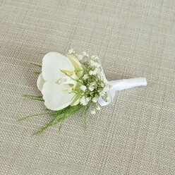 A refined white orchid boutonniere is captured lying gently on a pale beige, woven fabric background that suggests natural linen, its subtle texture giving the scene a calm, understated elegance often associated with intimate Epping weddings. At the centre, a flawless Phalaenopsis orchid bloom opens in serene symmetry, the petals smooth and velvety, curving outward in pure white. Within its centre, a whisper of pale yellow glows from the throat of the flower, offering a soft warmth that feels both romantic and tranquil, like a quiet stroll along the paths near Epping Forest. Around the orchid, clusters of fine baby's breath create an airy, mist-like halo; each tiny white blossom on its slender stem adds gentle volume without overpowering the central bloom. Delicate strands of light, feathery greenery weave between the Gypsophila, introducing refreshing splashes of soft green that hint at freshly cut stems and the subtle scent of foliage in a local florist's studio. The stems are neat and compact, bound with a crisp white satin ribbon that has a slight sheen, catching the light with a gentle glow. A small, tidy bow sits close to the base of the orchid, visually balancing the arrangement and indicating it is ready to be pinned to a groom's or groomsman's jacket. The overall effect is one of quiet sophistication and emotional clarity, suited to a wedding ceremony in Epping or nearby Loughton, where each floral detail is chosen to symbolise purity, commitment, and enduring love. The boutonniere appears freshly crafted, as if prepared the same morning by a skilled florist, ready for same-day wedding delivery across the local area.