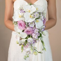 This captivating image conjures the delicate anticipation of a wedding morning in Epping, where a bride, viewed from mid-waist upward, holds a breathtaking bridal bouquet close to her heart. The neutral tan background is softly blurred, reminiscent of the golden hour over Epping Forest, allowing the arrangement to shine as the centrepiece. The bouquet cascades gracefully, shaped in an elegant teardrop that spills luxuriously downwards, much like the flow of her strapless gown with its intricate lace bodice glimmering at the edges of the frame. Pristine white phalaenopsis orchids reign at the forefront, their broad, flawless petals and sunny yellow centres exuding a sense of serene beauty. These are complemented by lush soft pink garden roses, velvety and fragrant, inviting associations with rambling roses near local Epping cottages. Wisps of lavender and pale purple stock flowers are woven delicately throughout, lending subtle spikes of texture and the fresh scent of a spring garden. Finely ruffled lisianthus, some edged prettily in deep amethyst, and airy sprigs of white baby's breath float amidst sparse green leaves, bringing volume without overwhelming the design. Together, the flowers create a visual and sensory symphony, embodying understated luxury perfect for intimate ceremonies in Epping churches or rustic venues. The image captures not only the rich craftsmanship of an experienced Epping florist, but also the emotional resonance-a bouquet that gently cradles the dreams and hopes of a romantic wedding celebration.