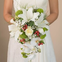 As the soft afternoon sun filters through ancient oaks in the heart of Epping, a bride holds an extraordinary cascading bouquet, expertly crafted with care and imagination. The striking display is defined by exquisite, snow-white lilies-each bloom unfurling in star-like splendor-accented by faint freckles of russet along the inner petals, creating a sense of rich depth and nuanced artistry. At the center, a classic creamy white rose curls inward, offering a focal point of serenity and traditional beauty that feels right at home in the timeless charm of Epping's countryside chapels. Sprigs of delicate white gypsophila add a cloud-like softness, drifting between lively tufts of lime-green pompom chrysanthemums that inject a joyful burst of color, reminiscent of wild meadows along Bower Hill. Vivid clusters of red Hypericum berries emerge like shining gems, offering a playful contrast to the bouquet's serene whites and verdant greens. Trailing elegantly, several large white Phalaenopsis orchids flow downward, their sculptural petals adding a sense of gracious movement and modern luxury-a nod to the sophisticated tastes of today's Epping brides. The hands of the bride, enveloped in an intricately detailed lace gown, cradle the bouquet with quiet anticipation. Soft, grass-like greenery and select leaves frame the entire arrangement, infusing it with a natural energy that reflects the vibrant spirit of Essex celebrations. This bouquet, alive with textural storytelling and local flair, radiates a sense of celebration and shared dreams, making it a perfect emblem for weddings throughout Epping and its charming surrounding hamlets.