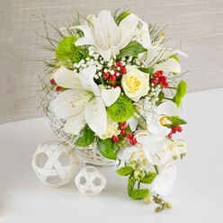 On a clean white tabletop, a lavish floral arrangement in a round white wicker basket commands attention, set against a softly textured beige background that feels like calm, filtered light in an Epping sitting room. At the heart of the design are several striking white lilies, some fully open with wide trumpet petals and delicately freckled throats, others poised to bloom, giving the bouquet a sense of unfolding promise. Nestled just above centre is a single creamy ivory rose, its petals spiralling in tight, velvety layers that suggest tenderness and new beginnings, ideal for a wedding breakfast at a local Epping venue. Flowing down from the right edge of the basket, a cascade of white phalaenopsis orchids creates a gentle waterfall, each orchid bloom with rounded, porcelain-like petals that evoke quiet luxury. Surrounding these focal flowers is an abundance of fresh greenery: fine, feathery fronds, broader glossy leaves, and bright green pom-pom blossoms that resemble miniature moss cushions, adding both vivid colour and tactile interest reminiscent of the nearby Epping Forest canopy. Small sprays of white baby's breath float like mist among the stems, while clusters of deep red berries punctuate the arrangement, bringing a subtle festive note without overpowering the soft palette. To the left, two decorative white woven balls, one larger and one smaller, rest on the table, echoing the basket's texture and introducing a sculptural, modern accent. The composition feels serene yet celebratory, as if prepared by an experienced local florist for a refined Epping wedding or anniversary dinner, where guests gather around the table and the flowers quietly set the tone of elegance and joy.