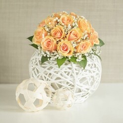 A lush dome of peach and apricot roses forms the heart of this elegant centerpiece, each rose carefully placed so the tightly furled petals reveal deeper, sunset-orange centers that gradually fade to softer, creamy edges. Threaded between the roses are delicate tufts of white baby's breath, their tiny blossoms creating a fine, misty halo that lightens the arrangement and gives it a dreamy, romantic character. Subtle touches of rich green foliage anchor the flowers, adding depth and keeping the composition from feeling overly sweet. All of this floral abundance rises from a striking spherical vase made from white woven strands, reminiscent of fine wicker or string art, its open latticework casting delicate shadows on the smooth white tabletop beneath. In front of the main vase, two smaller woven spheres lie playfully askew, echoing the same white material; one is larger and patterned in a way that hints at a modern, geometric football, while the other has a looser, more organic weave, both adding sculptural interest without detracting from the flowers. The background is a light beige or greige wall with a subtle vertical texture, quietly framing the scene so that the warm roses and intricate white textures remain the focus. The light is soft and even, as if filtering through a window overlooking Epping Forest, giving the arrangement a calm, welcoming glow. This image feels perfectly suited to illustrate a wedding table along Station Road or a stylish anniversary dinner at home in Epping, conveying the kind of careful craftsmanship and serene beauty local florists bring to intimate celebrations and same-day flower deliveries in the area.