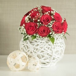 A rounded bouquet of crimson red roses forms the glowing centre of this photograph, each bloom at that perfect moment of openness when the petals are full, velvety, and richly layered. Around twelve to fifteen roses cluster tightly together, creating a smooth dome of colour that feels both passionate and composed, like a table arrangement ready to be placed at a small wedding reception in Epping. Sprays of fine white gypsophila, or baby's breath, drift between the roses, their tiny blossoms and slender stems creating airy pockets of light that contrast with the dense red petals. Occasional flecks of fresh green foliage are visible, tucked just beneath the heads of the roses, echoing the soft greens you might see strolling through Epping Forest on a late afternoon. The flowers are gathered into a distinctive white vase with a bulbous, rounded shape and an intricate open-weave design. Thin white strands crisscross and loop around each other, forming a basket-like lattice that feels modern yet handcrafted, much like the artisan pieces you might find in small independent shops off Epping High Street. The vase rests on a simple white surface, its clean lines giving a gallery-like setting to the arrangement. To the left, two off-white decorative spheres made from flat, woven strips sit casually on the same surface, one noticeably larger than the other, echoing the texture and shape of the vase while adding playful balance. The background is a softly blurred expanse of neutral beige, slightly textured like a fine fabric, allowing the roses and woven details to hold full attention. The scene conveys intimate romance and understated elegance, ideal for a special dinner, Valentine's surprise, or refined home décor in and around Epping.