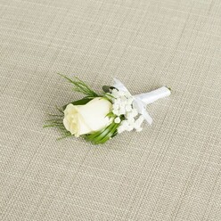 A refined white rose boutonniere is displayed at a diagonal across a neutral, linen-style beige background, as if carefully laid out on a dressing table just before a wedding in Epping. The central rosebud is a creamy white, its petals smooth and tightly coiled, with only the slightest unfurling at the edges to suggest a fresh bloom at its peak. The rose feels soft and velvety to the imagination, with a gentle ivory warmth that stands out clearly against the muted fabric. Surrounding the flower, a light spray of delicate white baby's breath forms a misty halo, each minuscule blossom resembling a dot of lace, giving the boutonniere a romantic, cloud-like texture. Tucked behind and beside the rose are slim, dark green leaves and a few fine, fern-like fronds of foliage, adding contrast and structure without overpowering the simple elegance of the design. These touches of greenery evoke the look of a hand-picked stem from a leafy path by Epping Forest, then refined by a florist's practiced hands. The stems are neatly aligned and wrapped in a narrow band of pure white satin ribbon, the surface smooth and lightly reflective under the soft, even lighting. Just below the floral head, the ribbon is fashioned into a dainty bow, its loops crisp and symmetrical, signalling care and craftsmanship suitable for a groom or groomsman. The fabric beneath has a gentle woven crosshatch pattern, like natural linen or raw silk, contributing to the understated, organic setting. The overall scene radiates calm joy and anticipation, making the boutonniere an ideal visual for discussing groom's accessories, same-day wedding flower delivery around Epping, or the art of creating traditional lapel flowers.