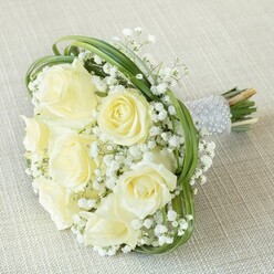 Laid gently upon a subtly textured beige cloth, this enchanting bouquet captures the timeless romance cherished in Epping ceremonies and celebrations. Centerpiece blooms of creamy white and butter-hued roses command attention, their softly curling petals revealing the delicate stages of new blooms to full, opulent flowers. Airy clouds of baby's breath are interlaced between each rose, giving the bouquet a lightness reminiscent of early mornings by the market square or the tranquil beauty found near Epping's High Street gardens. Sleek arcs of deep green foliage-likely bear grass gathered fresh from local sources-loop gracefully throughout, adding structure and modern flair that balances tradition with contemporary style. The bound stems, neatly fixed into a smooth handle, are wrapped in a silvery-grey ribbon and exquisitely finished with gleaming rows of pearl embellishments, suggesting both sophistication and heartfelt care. The tips of the natural stems peek out beneath, highlighting the freshness of the hand-tied arrangement. This bouquet, crafted for precious moments from weddings to anniversaries, radiates serenity and warmth-ready for same-day delivery to homes or venues around Epping, making every event shine with understated grace.