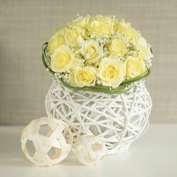 On a pristine white tabletop, a carefully crafted dome of pale yellow roses becomes the calm heart of the scene, each bloom at that perfect point of openness where the petals look velvety and full, like swirls of fresh cream. Dozens of roses are nestled tightly together so no gaps show, their buttery tones shifting from soft lemon at the centre to a warmer, creamy yellow at the outer petals. Fine sprays of white baby's breath weave through the roses, forming a delicate mist that softens the arrangement and introduces tiny points of brightness, as if catching the same gentle daylight you might find drifting through an Epping townhouse window. A slim band of vivid green foliage wraps around the base, creating a neat visual boundary between the blooms and the vessel. The flowers sit in a sculptural white woven vase, its open rattan-like strands looping and crisscrossing to form a rounded shape that narrows slightly in the middle before curving out again. Light passes through the gaps, creating subtle patterns of shadow on the gleaming surface below. To one side are two decorative woven balls, their light timber tones and airy construction echoing the texture of the vase and adding a touch of organic, Nordic-style design. The softly blurred, neutral background keeps distractions away, allowing the eye to rest fully on the flowers and their intricate home. This image feels perfect for illustrating wedding table arrangements for receptions around Epping, Theydon Bois, or Loughton, where couples want understated luxury and a peaceful, contemporary look.
