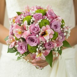 In Epping's gentle sunlight, a bride tenderly holds a bouquet where colour, texture, and form merge in harmonious celebration. Mauve and pale pink roses, some petals dappled with dew, swirl at the bouquet's heart, offering a nostalgic nod to cherished gardens bordering local lanes like Bell Common and Ivy Chimneys. A lively ring of lime green button chrysanthemums enlivens the design, their playful, rounded shapes adding a crisp energy quintessential to modern bridal trends. Nestled between are delicate pink alstroemeria, their streaked petals and subtle tropical scent a whisper of the world's wider beauty, freshly sourced from regional suppliers. Throughout the bouquet, ethereal clouds of white baby's breath impart a dreamy softness, tying the elements together like memories woven through a celebration. Dark glossy foliage frames the composition, providing structure and accentuating each hue. The bride's hands, softened by the textured lace of her dress, cradle the florals as if holding whispered promises of Epping's fresh beginnings and lifelong journeys. Every detail, lovingly arranged by Flowers Epping, speaks of skill and devotion-creating a bouquet that feels as personal as a stroll through the town's historic High Street. It's an arrangement that captures both time-honored romance and the spirited pulse of modern weddings celebrated in Epping and its lush surroundings.