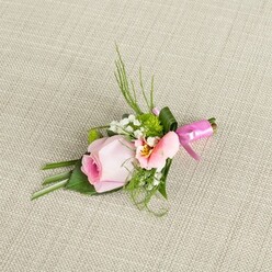 This delicate boutonniere, crafted with enduring care near the leafy edges of Epping, offers a radiant harmony of pastel hues and graceful form. At its core lies a bashful, soft pink rose-its petals rolled tightly in a gentle arc, the colour deepening at its heart before blushing into pale tones as each layer softly unfurls. Alongside it, a lighter, peach-tinted flower, possibly Alstroemeria, reveals subtle sunlit yellow and intricate brushstroke markings, lending delicate complexity reminiscent of early morning strolls in Epping's market square. Wisps of baby's breath float throughout the piece, appearing as if kissed by dew, while vivid green spheres, like miniature chrysanthemums, nestle between blossoms, each one echoing the vibrant green spaces that surround the town. Lush, textural greenery gathers below: long, slender blades evoke the familiar grasses along Stonards Hill, while broader leaves, some elegantly folded, set a sturdy foundation. The entire arrangement is united by a shimmering fuchsia satin ribbon, tied with precision in a neat, celebratory bow, its sheen vibrant beneath soft, natural light. Resting on a neutral, coarse-textured beige linen reminiscent of artisan textiles found in local shops, this boutonniere is designed for moments of romance and milestones: a groom's lapel at a wedding in Theydon Bois, or a touch of grace at a special celebration nearby. Its palette, freshness, and gentle fragrance embody the artistry of Epping floristry and the uniqueness of every special occasion.