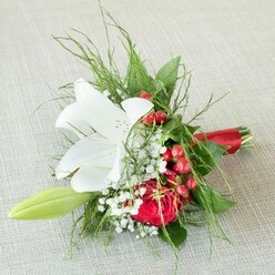 Laid gracefully atop a subtle, linen-textured surface that echoes Epping's rustic elegance, this small bridal bouquet emanates refined festivity. The focal white lily, pristine as morning dew upon Epping Common, displays broad petals freckled with reddish-brown, its golden anthers glowing with vibrant life. To its side, a deeply saturated red rose unfurls, symbolizing enduring affection, its plush petals spiraling to the center-just as community ties coil strongly through local celebrations. Sprigs of snowy white gypsophila weave a magical softness between the structured florals, their meadowlike quality familiar to those who walk Epping's garden paths. Festive red berries-bold and lustrous-wink like treasures discovered in local hedgerows, providing a joyful, seasonal spark. A fresh lily bud points upward, foretelling future blossoms and continued happiness, while layers of textured green ferns and smooth, broad leaves embrace the floral core with woodland tranquility. Everything is finished with a smart red satin ribbon wrap, ensuring the bouquet is ready for cherished moments-be it an intimate wedding at a local Epping venue or a thoughtful gift for delivery to nearby Market Street. This arrangement unites classic romance and local natural beauty, shining with meticulous care and tenderness from Flowers Epping's skilled hands.