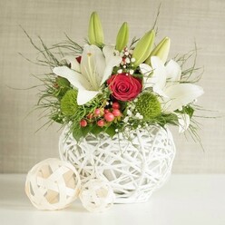 A sophisticated floral centerpiece is displayed as if ready to grace a wedding table in one of Epping's charming country venues, resting on a bright white surface against a gently textured neutral wall. The eye is immediately drawn to the distinctive round vase, crafted from matte white strands that weave and loop into an airy, open lattice, evoking handwoven baskets found at local markets near Epping Forest. Overflowing from this sculptural container is a carefully composed bouquet of white lilies, rich red roses, and textured greenery. Several lilies are fully open, their broad, creamy petals curving gracefully back to reveal soft freckles and warm golden stamens, while other buds stand tall and closed, signaling freshness and longevity. Between the lilies, deep scarlet roses with tightly coiled centers introduce a note of romance, each velvety petal catching the gentle light. Bright green pom-pom flowers, likely green dianthus or chrysanthemums, punctuate the design with playful, spherical shapes and a lively splash of colour. Fine sprays of baby's breath drift through the arrangement, their miniature white blossoms creating a light, cloud-like veil around the bolder blooms. Small clusters of red berries, reminiscent of seasonal hedgerows along the lanes out towards Theydon Bois, add a subtle festive accent that suits both winter weddings and special occasion dinners. Wispy, dark green fern-like foliage and broader leaves cascade slightly over the rim of the vase, giving the piece a soft, natural silhouette. To the left, two decorative wicker-style balls in pale beige echo the woven design of the vase, lending a harmonious finishing touch. This composition feels calm, romantic, and meticulously crafted, the sort of centerpiece an Epping-based florist would design for an elegant local celebration.