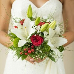 In the heart of Epping, a bride is captured in a moment of luminous grace, clad in a subtly textured white gown as she holds a lavish bouquet against her waist. The arrangement, designed with an artist's intuition, draws the eye with grand white oriental lilies-some majestic and open, showing off golden stamens, others as pale buds hinting at future beauty. Between these lilies, deep red roses curl in silken spirals, sending a quiet message of fervor and enduring love. Bright red berries offer playful punctuation, their glossy skins gleaming alongside soft tufts of baby's breath, which create clouds of lightness around the focal blooms. Clusters of fresh green spheres and an array of textured foliage-from the finely feathered to the broad and robust-evoke walks through the greenways of Epping and the markets of the High Street. Held gently in the bride's hands, the bouquet mirrors the careful thought and heartfelt emotion that Flowers Epping brings to every wedding. The soft background, illuminated by natural daylight, ensures every petal's detail stands out, framing a bouquet that isn't just a decoration but a part of the story, woven into the memories of local couples tying the knot in Epping.
