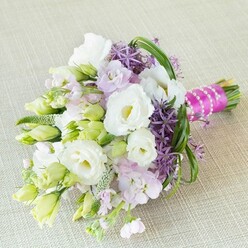 An elegant bridal bouquet is artfully presented atop a soft, neutral-toned backdrop, its hues and textures inspired by the gentle countryside that surrounds Epping. The centrepiece features lush white Lisianthus blooms-unfurling in creamy, ruffled splendour-flanked by tightly coiled buds that promise freshness and new life. Scattered throughout are clusters of pale lavender-pink stocks and intricate, deep purple allium, their starburst silhouettes offering dramatic shape and sense of movement reflective of an early morning stroll through Epping Forest. Slim, arching blades of vibrant green foliage and feathery textured fillers echo the natural abundance found near the High Street flower stalls, intertwining with lime-green buds to create depth and lively energy. Binding the bouquet is a vivid fuchsia ribbon, elegantly wrapped and cross-laced with pearl strands that shimmer softly, imparting a refined touch reminiscent of timeless nuptial traditions cherished by local couples. Bathed in gentle light, the pastel palette glows, invoking the serene optimism and anticipation that fills a wedding morning across Epping's historic churches and elegant venues. This bouquet is carefully crafted not simply to be admired, but held close-an artistic expression of unity, romance, and enduring connection, delivered with the expertise and personal care for which local florists are known.