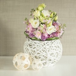 A refined floral composition is captured as though it were the heart of a bright Epping townhouse, resting on a spotless white table bathed in soft, diffused daylight. At the centre, a rounded white vase made from an intricate weave of painted rattan or slender twigs cradles a generous bouquet that seems to billow upward like a gentle spring breeze rolling over Epping Forest. Creamy white and pale yellow lisianthus form the main body, their velvety, multi-layered petals curling delicately at the edges, giving the impression of movement even in stillness. Nestled among them, stems of stock in tender blush pink and muted lavender reach a little higher, their clustered blossoms adding both fragrance and a sculptural, vertical line that suggests the hand of an experienced florist. Around the base, clusters of small, vivid purple filler flowers build a lush, textured carpet, deepening the colour palette and drawing the eye inward, while a slim ring of glossy, dark green leaves frames the entire arrangement with quiet elegance. The vase itself feels both modern and organic, its open latticework allowing light to pass through and cast subtle shadows that dance across the white surface. To the left, two light wooden or rattan spheres echo the woven motif, the larger one slightly forward, the smaller tucked behind, like a pair of thoughtfully chosen accents on a sideboard along Theydon Road. Behind, a soft beige or greige wall with a faint vertical fabric texture provides a calm, neutral backdrop. The overall atmosphere is tranquil and intimate, ideal as a centrepiece for a small wedding breakfast or an anniversary supper at home, the sort of bespoke design Flowers Epping might create for local families seeking something understated yet memorable.